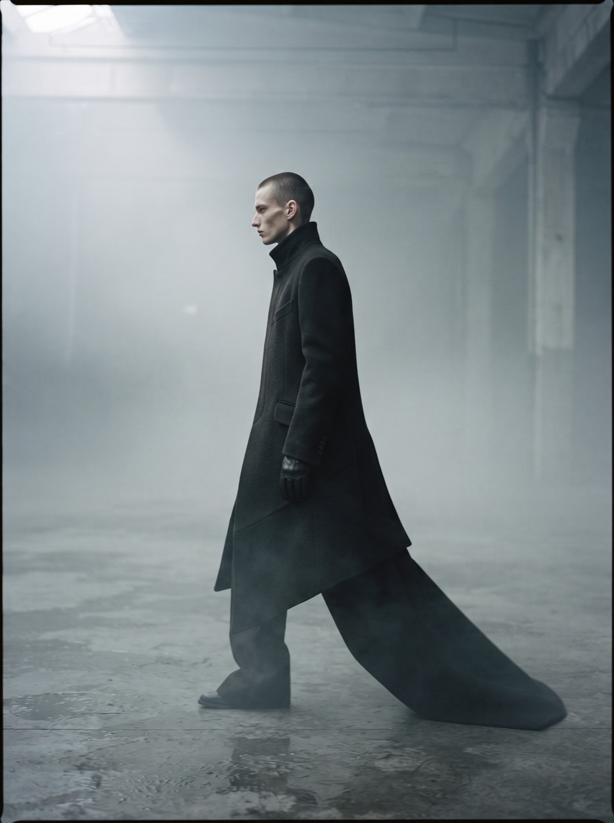A model in profile walking through fog in an off-black sculptural coat with a long trailing asymmetric hem.