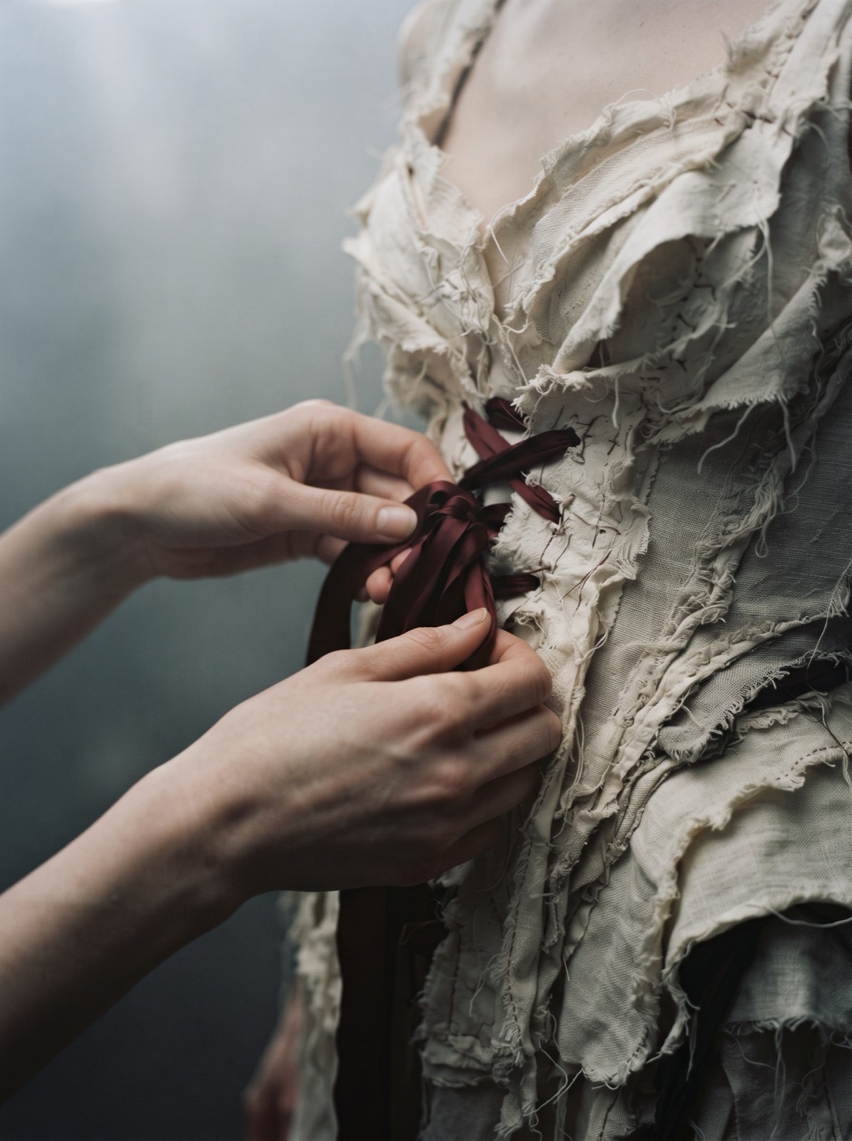 Close detail of hands adjusting a bone-white layered bodice with raw edges and a single oxblood ribbon.
