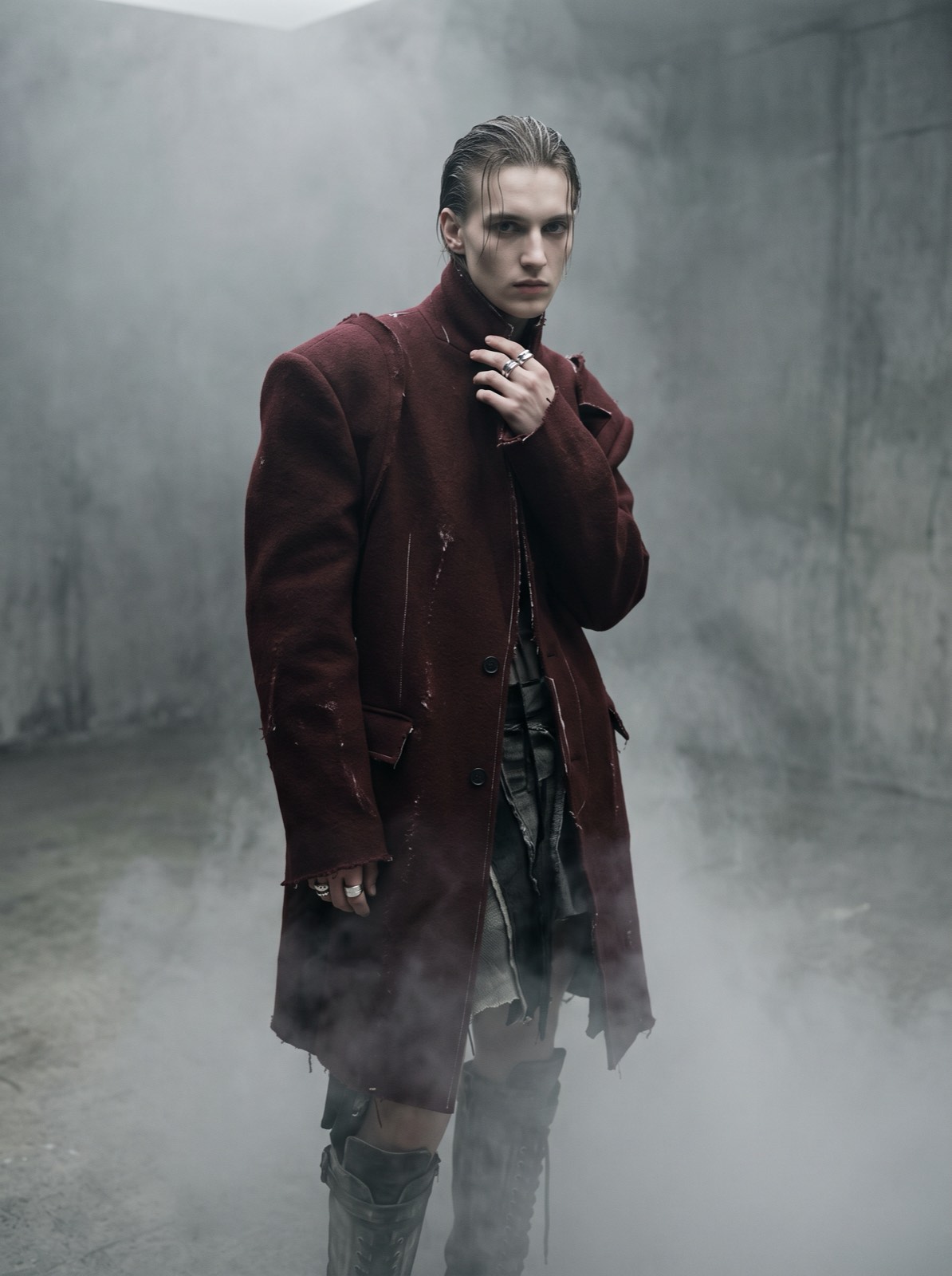 Three-quarter portrait of a model in a deconstructed oxblood wool overcoat with sculpted shoulders, against grey fog.